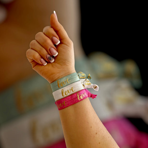 Diese elastischen Armbänder tragen den Schriftzug Love und sind in Salbei, Weiß und Pink/- Gold abgebildet.
